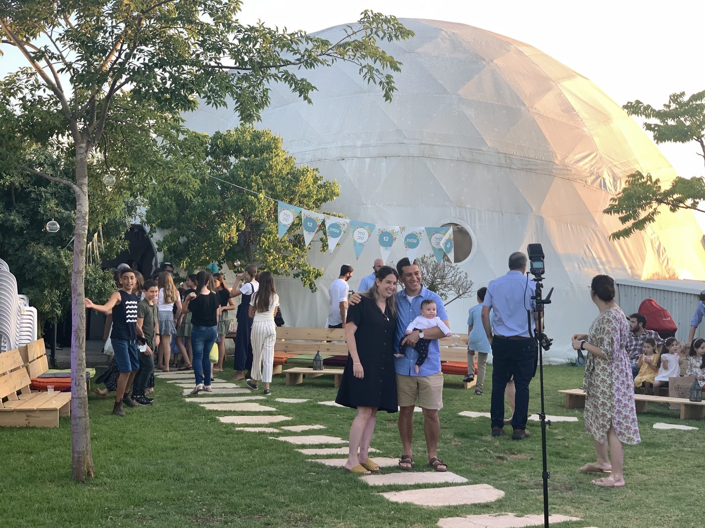 An evening gathering beneath the great dome at Havat HaChaim, with bunting, benches, and families on the grass
