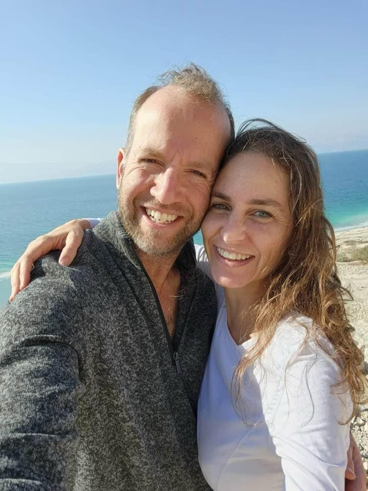 Tai and his wife Shira cheek to cheek, smiling, with the turquoise water of the Dead Sea behind them
